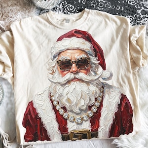 Może przedstawiać: Kremowy t-shirt z grafiką Świętego Mikołaja w okularach przeciwsłonecznych w panterkę i dużym, masywnym złotym łańcuchu. Grafika wykonana jest w stylu grubych, teksturowanych pociągnięć pędzla, przedstawiająca Mikołaja w czerwonym stroju.