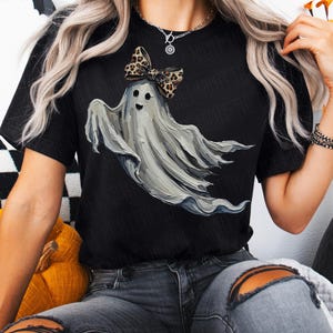 May include: Black t-shirt with a ghost illustration. The ghost is grey and wears a leopard print bow. The design is a fun take on Halloween themes.