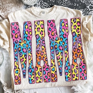 May include: A cream-colored short-sleeve t-shirt featuring the word MAMA printed in large block letters. The letters have a distressed, multi-colored leopard print pattern in shades of pink, yellow, blue, and orange against the fabric.
