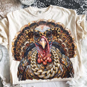 May include: A cream-colored t-shirt featuring a graphic print of a turkey wearing leopard-print sunglasses and a thick, gold-toned chain necklace with embedded crystals. The turkey is rendered in a textured, painterly style with brown, tan, and red tones.