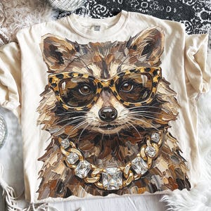 May include: Cream-colored t-shirt featuring a detailed illustration of a raccoon wearing leopard print glasses and a gold chain with large square gems. The raccoon's fur is rendered in shades of brown and tan.