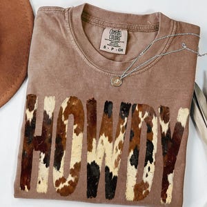 May include: A brown t-shirt with the word "HOWDY" in a cowhide pattern. The shirt has a crew neck and short sleeves. A silver necklace with a pendant is draped across the shirt. A brown hat is visible in the upper left corner.