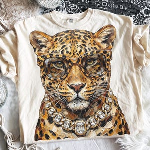 May include: A cream-coloured t-shirt with a detailed illustration of a leopard wearing glasses and a diamond necklace. The leopard has a spotted coat and is centered on the shirt. The shirt is made of a soft material.