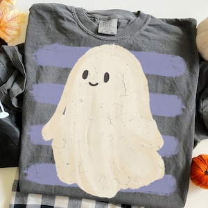 May include: A charcoal gray crew neck t-shirt featuring a large, cream-colored ghost graphic with a simple smiling face. The ghost is layered over horizontal, distressed light purple brushstroke stripes. The shirt has a tag reading Comfort Colors.
