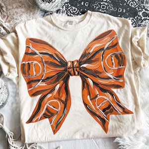 May include: Cream-colored t-shirt featuring a large, orange bow design with black and white accents. The bow has a basketball court design. The shirt is made of a soft material.