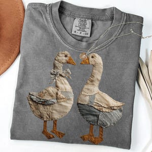 May include: Gray Comfort Colors t-shirt featuring two appliqued geese. One goose has a bow tie, the other has overalls. A silver necklace with a small pendant is draped across the shirt. The shirt has a small tag that says "Comfort Colors".