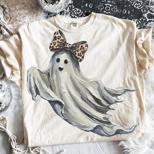 May include: Cream-colored t-shirt featuring a whimsical ghost illustration. The ghost is painted in shades of gray and white, wearing a leopard print bow. The design is centered on the shirt, creating a playful and unique look.