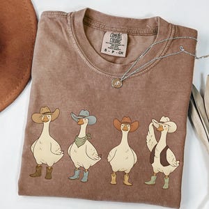 May include: A brown Comfort Colors t-shirt with a graphic of four geese dressed as cowboys, wearing hats and boots. One goose has a vest and another a bandana. A silver necklace is draped across the shirt. The shirt is a small size.