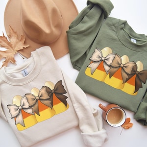 May include: Two sweatshirts, one cream and one olive green, each featuring a candy corn design with a bow. A brown hat and a fall leaf are also visible. The sweatshirts are likely for the fall or Halloween season.