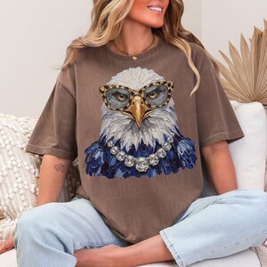 May include: A brown short-sleeve t-shirt featuring a graphic of a bald eagle wearing leopard print glasses and a large crystal-style necklace. The eagle has white head feathers and dark blue chest feathers, rendered in a textured, painterly style.