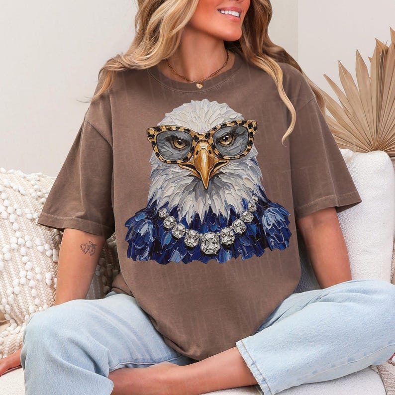 May include: A brown short-sleeve t-shirt featuring a graphic of a bald eagle wearing leopard print glasses and a large crystal-style necklace. The eagle has white head feathers and dark blue chest feathers, rendered in a textured, painterly style.
