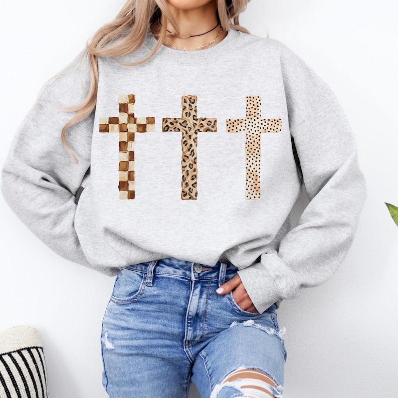 May include: A light gray sweatshirt featuring three crosses with different patterns. The crosses have a checkered, leopard print, and dotted design. The sweatshirt is paired with blue jeans.