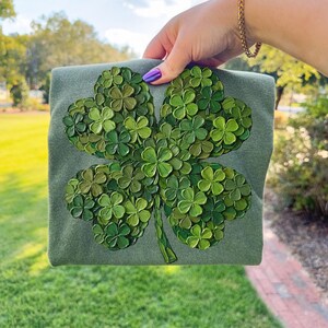 May include: A green sweatshirt featuring a large, four-leaf clover design. The clover is composed of many smaller, textured green clovers, creating a 3D effect. The sweatshirt is held up against a blurred outdoor background.