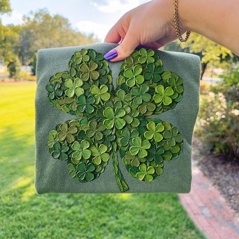 May include: A green sweatshirt featuring a large, four-leaf clover design. The clover is composed of many smaller, textured green clovers, creating a 3D effect. The sweatshirt is held up against a blurred outdoor background.