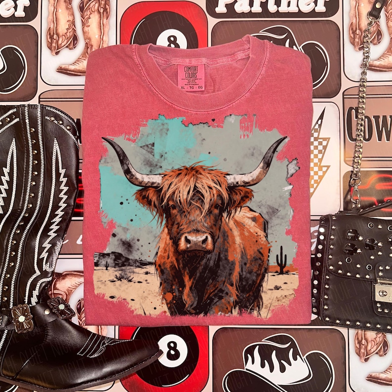 May include: A red t-shirt with a Highland cow illustration in a watercolour style. The design includes a desert scene with cacti and a turquoise and grey backdrop. The shirt has a Comfort Colors label.