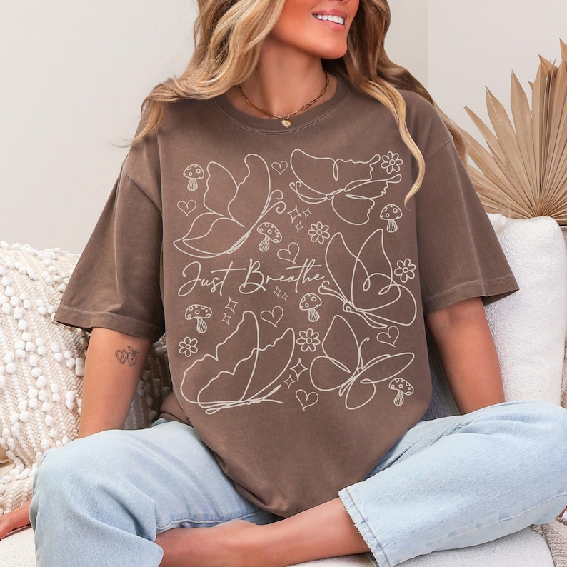 Puede incluir: Camiseta marr&oacute;n con un dise&ntilde;o de mariposas y flores blancas, y el texto "Just Breathe". El dise&ntilde;o incluye mariposas, corazones, flores y setas. La camiseta es de corte holgado.
