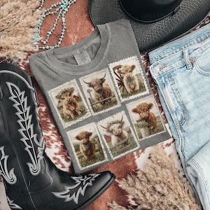 May include: A gray t-shirt featuring six postage stamp designs with illustrations of Highland cows. The shirt is laid flat with a black cowboy boot, blue jeans, a turquoise necklace, and a black hat.