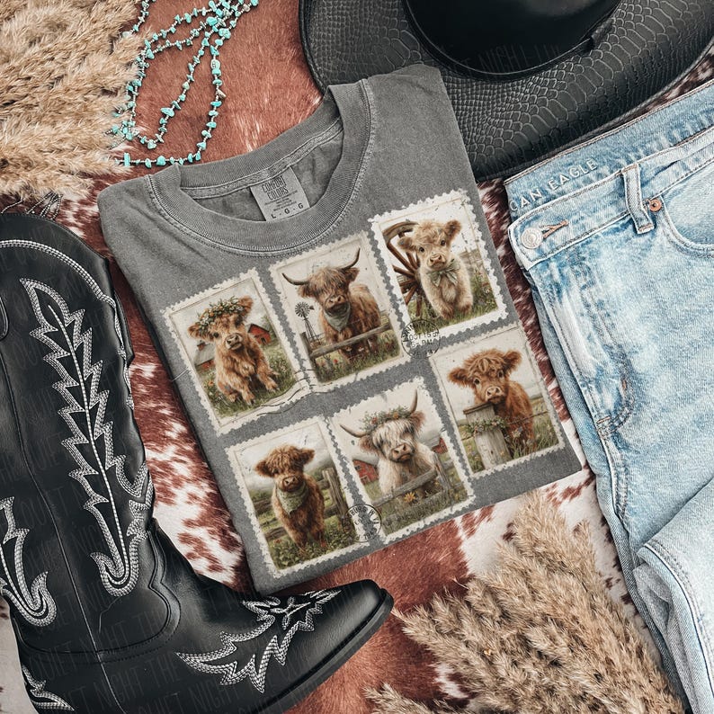 May include: A gray t-shirt featuring six postage stamp designs with illustrations of Highland cows. The shirt is laid flat with a black cowboy boot, blue jeans, a turquoise necklace, and a black hat.