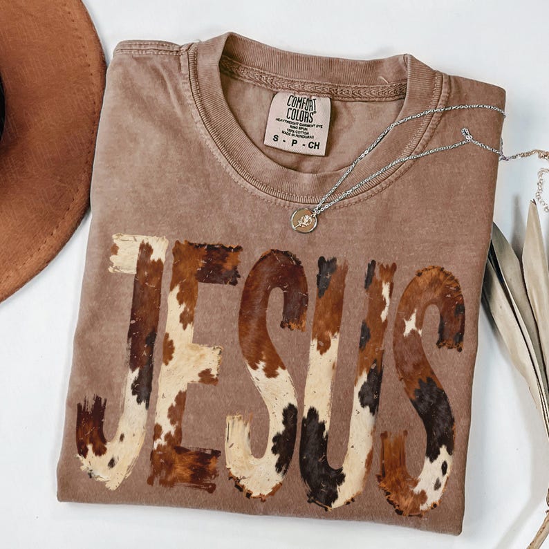 May include: A brown Comfort Colors t-shirt with the word "JESUS" in a cowhide pattern. A silver necklace with a pendant rests on the shirt. A brown hat is partially visible in the upper left corner.