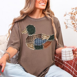 May include: A brown short-sleeve t-shirt featuring a large duck graphic on the chest. The duck is composed of various fabric patches with different patterns, including plaid, floral, and solid textures, all with frayed, raw edges.