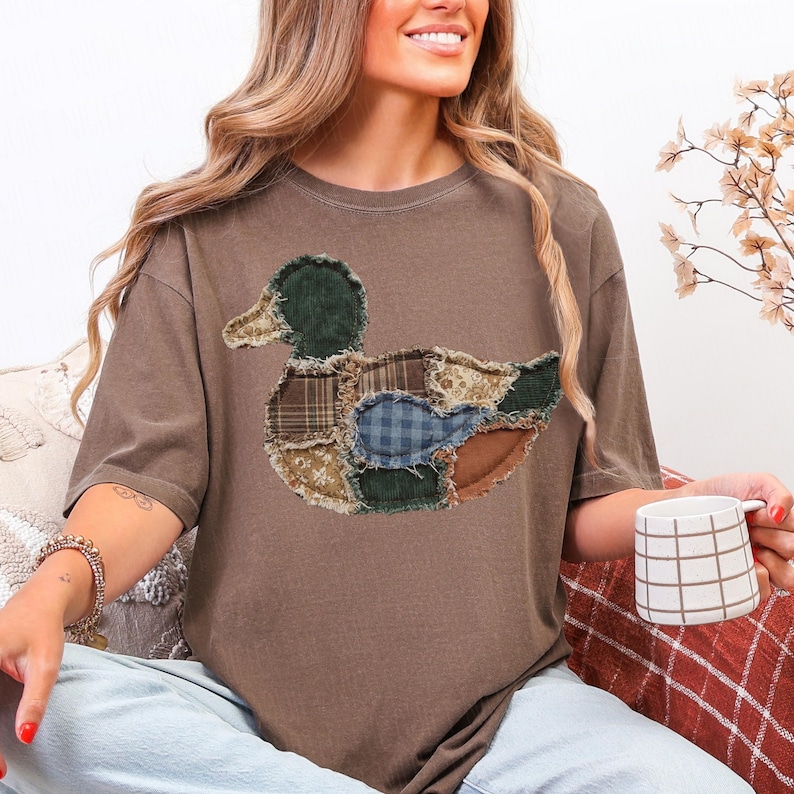 May include: A brown short-sleeve t-shirt featuring a large duck graphic on the chest. The duck is composed of various fabric patches with different patterns, including plaid, floral, and solid textures, all with frayed, raw edges.