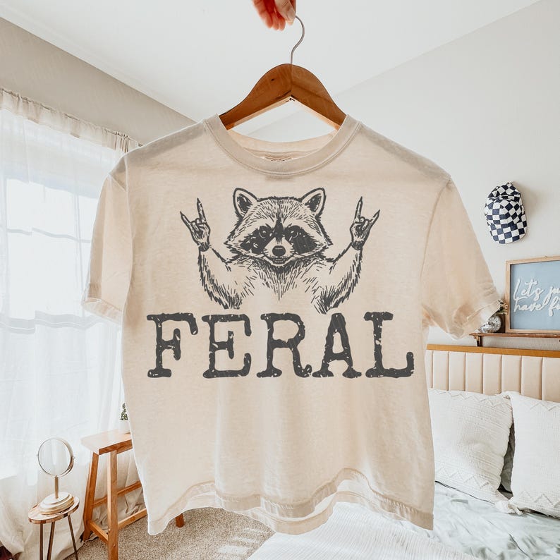 May include: Cream-colored t-shirt with a graphic of a raccoon making a rock-and-roll hand gesture. The word "FERAL" is printed in bold, dark gray letters below the raccoon illustration. The shirt is hanging on a wooden hanger.