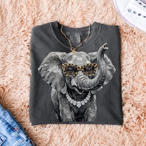 May include: A charcoal gray crewneck t-shirt featuring a graphic of an elephant wearing leopard print glasses and a crystal necklace. The elephant illustration has a textured, painterly style with visible brushstrokes in shades of gray.