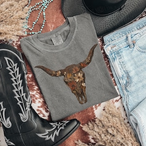 May include: A gray t-shirt featuring a floral bull skull design, paired with blue jeans, a black cowboy hat, and a black cowboy boot with white stitching. The items are arranged on a cowhide-patterned surface, with turquoise beads and dried grass.