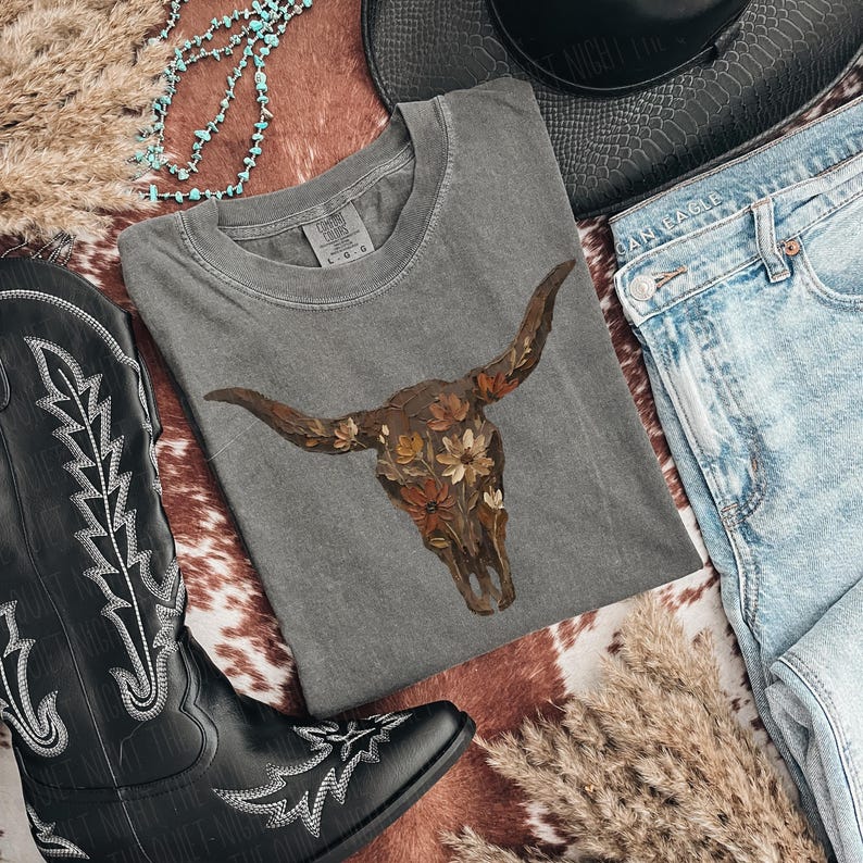 May include: A gray t-shirt featuring a floral bull skull design, paired with blue jeans, a black cowboy hat, and a black cowboy boot with white stitching. The items are arranged on a cowhide-patterned surface, with turquoise beads and dried grass.