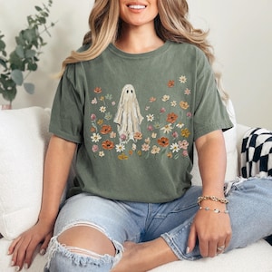 Może przedstawiać: Oliwkowozielony t-shirt z kr&oacute;tkim rękawem z centralną grafiką przedstawiającą białego ducha otoczonego r&oacute;żnorodnymi kolorowymi polnymi kwiatami w odcieniach pomarańczu, ż&oacute;łci i r&oacute;żu. Koszulka ma okrągły dekolt i luźny, swobodny kr&oacute;j.