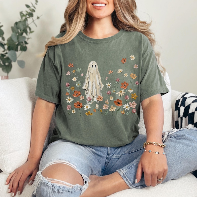 Może przedstawiać: Oliwkowozielony t-shirt z kr&oacute;tkim rękawem z centralną grafiką przedstawiającą białego ducha otoczonego r&oacute;żnorodnymi kolorowymi polnymi kwiatami w odcieniach pomarańczu, ż&oacute;łci i r&oacute;żu. Koszulka ma okrągły dekolt i luźny, swobodny kr&oacute;j.