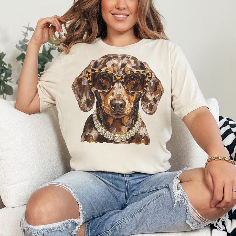 May include: A cream-colored short-sleeve t-shirt featuring a graphic print of a dapple dachshund dog wearing leopard print glasses and a gold-toned chain necklace. The dog has brown and tan mottled fur and is centered on the front of the shirt.