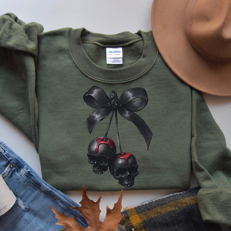 May include: An olive green sweatshirt with a graphic of two black skull cherries with red drips, tied with a black bow. The crew neck sweatshirt has a tag on the collar.