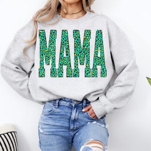 K&ouml;nnte beinhalten: Ein hellgraues Sweatshirt mit Rundhalsausschnitt und dem Wort MAMA in gro&szlig;en Blockbuchstaben. Die Buchstaben haben ein leuchtendes t&uuml;rkisfarbenes und gelbes Leopardenmuster. Das Sweatshirt hat lange &Auml;rmel sowie gerippte B&uuml;ndchen am Hals und Saum.