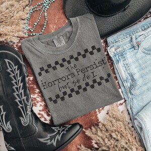 May include: A gray t-shirt with the text "The Horrors Persist but so do I" in a distressed font. The shirt is laid flat with a pair of blue jeans, a black cowboy boot, a black hat, and turquoise beaded necklace.