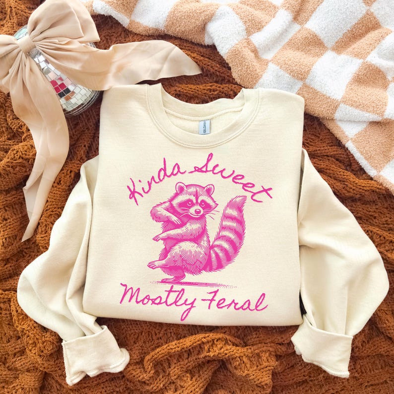 May include: Cream-colored sweatshirt with a pink raccoon graphic and the text "Kinda Sweet, Mostly Feral." The raccoon is standing on its hind legs. The sweatshirt has long sleeves and a crew neck.
