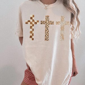 May include: Cream-colored t-shirt featuring three crosses with unique patterns. The crosses include a checkered design, a leopard print, and a dotted pattern. The shirt is a relaxed fit.