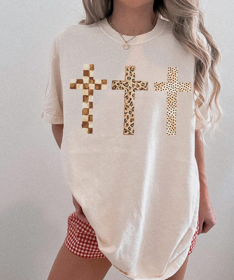 May include: Cream-colored t-shirt featuring three crosses with unique patterns. The crosses include a checkered design, a leopard print, and a dotted pattern. The shirt is a relaxed fit.