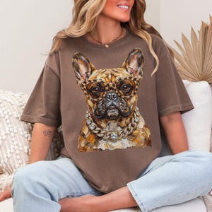 May include: Brown t-shirt featuring a detailed illustration of a French Bulldog wearing leopard print glasses and a diamond chain. The dog's fur is a mix of brown and tan. The shirt is a solid brown color.