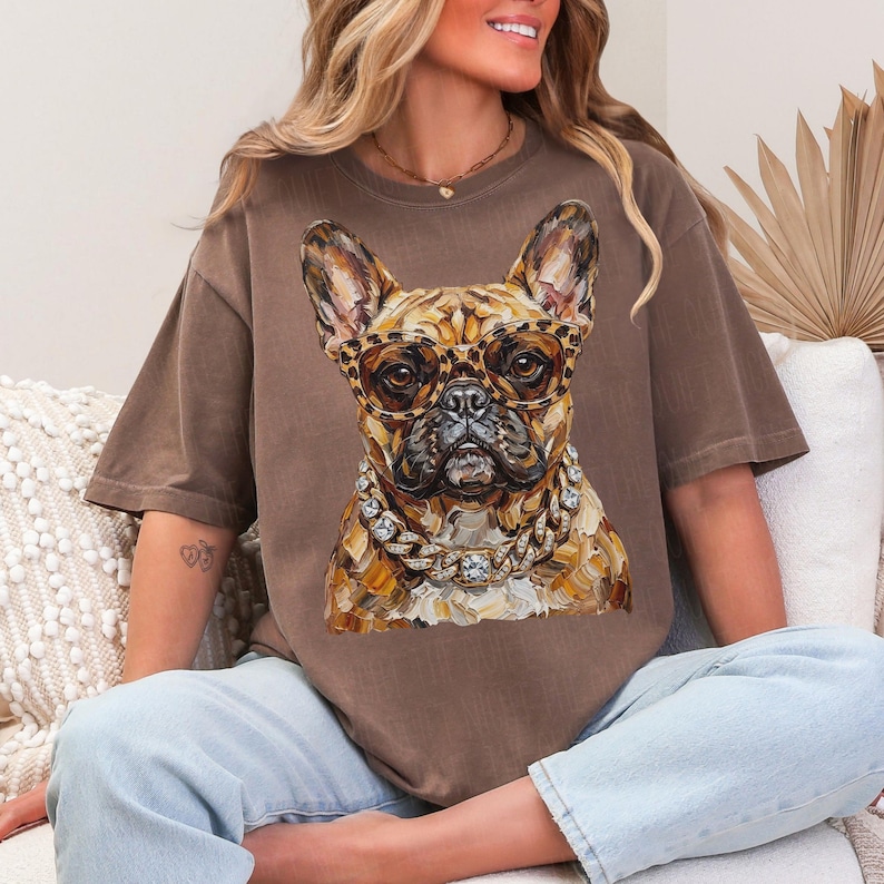 May include: Brown t-shirt featuring a detailed illustration of a French Bulldog wearing leopard print glasses and a diamond chain. The dog's fur is a mix of brown and tan. The shirt is a solid brown color.
