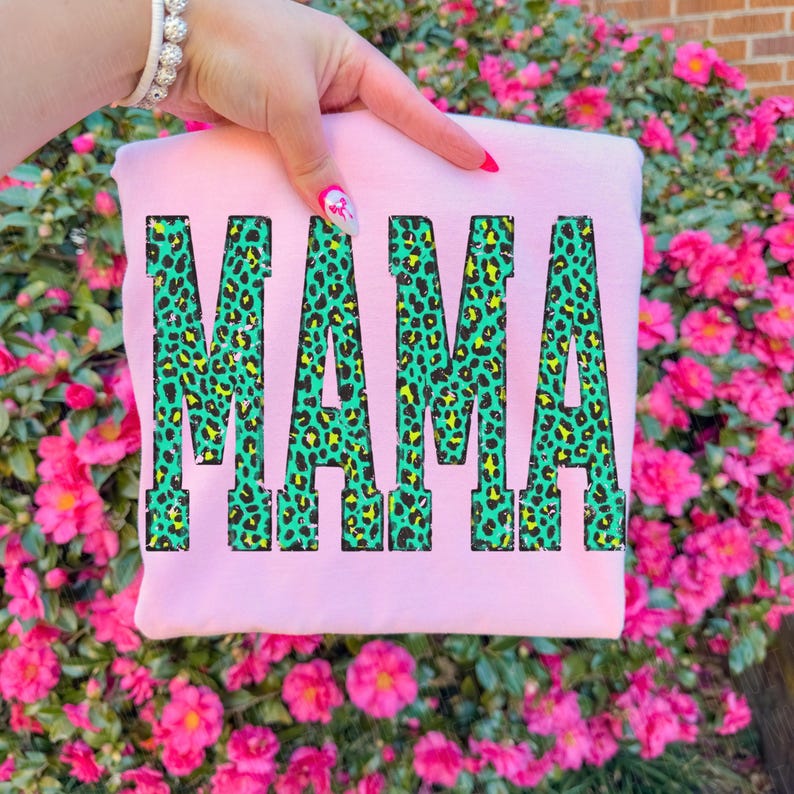 K&ouml;nnte beinhalten: Ein hellrosa Sweatshirt mit dem Wort MAMA in gro&szlig;en Blockbuchstaben. Die Buchstaben haben ein leuchtend gr&uuml;nes und schwarzes Leopardenmuster. Das Kleidungsst&uuml;ck wird vor einem Hintergrund aus leuchtend rosa Blumen pr&auml;sentiert.