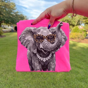 May include: A bright neon pink t-shirt featuring a graphic print of a grey elephant with textured brushstroke details. The elephant wears leopard print glasses and a silver-toned necklace with clear, faceted stones around its neck.