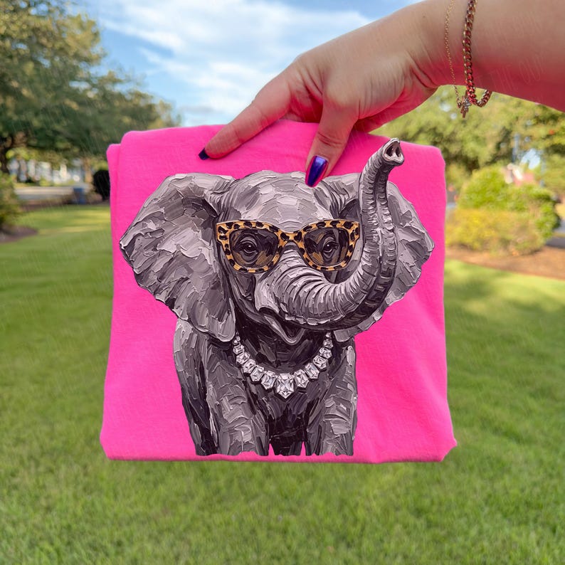 May include: A bright neon pink t-shirt featuring a graphic print of a grey elephant with textured brushstroke details. The elephant wears leopard print glasses and a silver-toned necklace with clear, faceted stones around its neck.
