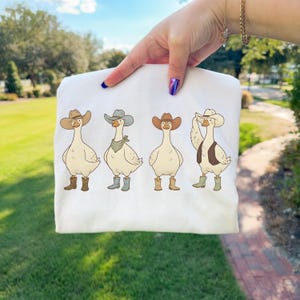 May include: White sweatshirt featuring a cartoon design of four geese dressed as cowboys. Each goose wears a cowboy hat, boots, and some have bandanas or vests. The geese are in various poses, adding a playful touch to the design.