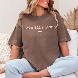 May include: A brown t-shirt with the text "love like jesus" and a cross graphic. The shirt has short sleeves and a relaxed fit. The model is wearing light blue jeans.