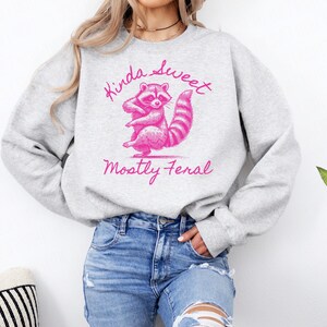 May include: A light gray sweatshirt with a pink graphic of a raccoon and the words "Kinda Sweet, Mostly Feral". The raccoon is standing on its hind legs with a striped tail. The sweatshirt is a casual style.