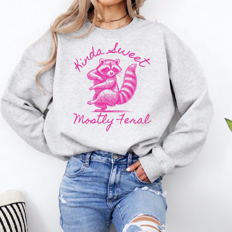 May include: A light gray sweatshirt with a pink graphic of a raccoon and the words "Kinda Sweet, Mostly Feral". The raccoon is standing on its hind legs with a striped tail. The sweatshirt is a casual style.