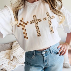 May include: White t-shirt featuring three crosses with unique patterns. The crosses have a checkered, leopard print, and polka dot design. The shirt is paired with light blue jeans.