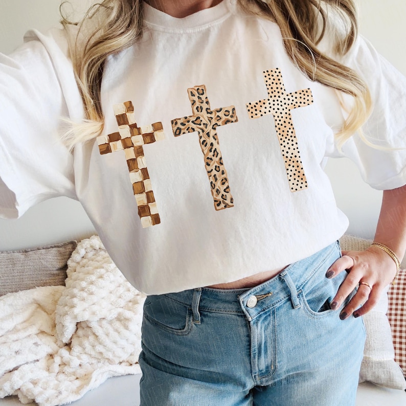 May include: White t-shirt featuring three crosses with unique patterns. The crosses have a checkered, leopard print, and polka dot design. The shirt is paired with light blue jeans.