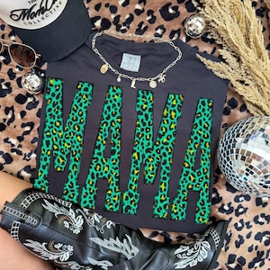 K&ouml;nnte beinhalten: Ein schwarzes T-Shirt mit dem Wort MAMA in gro&szlig;en Blockbuchstaben in einem leuchtend gr&uuml;nen und schwarzen Leopardenmuster. Das Shirt ist mit einer goldfarbenen Kette, einer schwarzen Baseballkappe und einem schwarzen Lederstiefel mit wei&szlig;er Stickerei kombiniert.